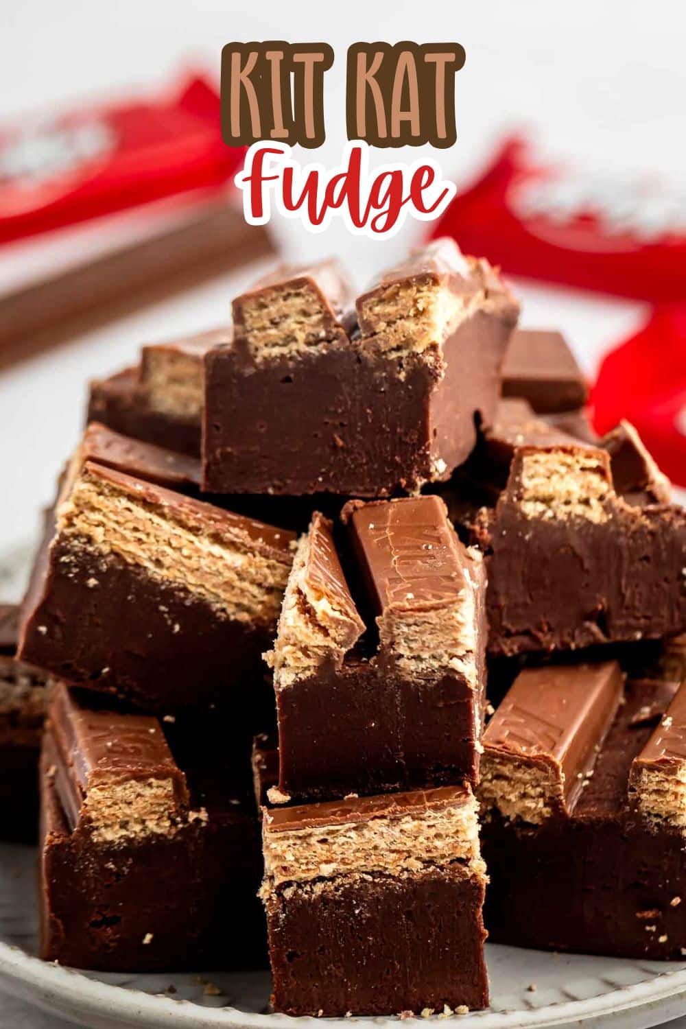 A plate stacked with square pieces of chocolate fudge layered with Kit Kat bars, showing crispy wafer layers inside. Kit Kat wrappers are blurred in the background. Text at the top reads Kit Kat Fudge.
