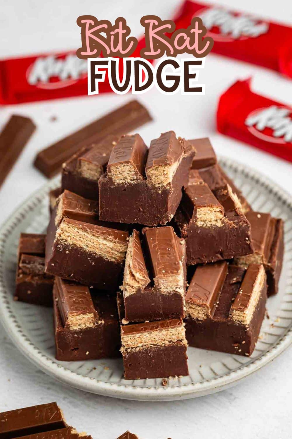 A plate stacked with pieces of chocolate fudge containing Kit Kat wafer bars, with red Kit Kat wrappers and extra chocolate pieces in the background. The text Kit Kat Fudge appears at the top of the image.