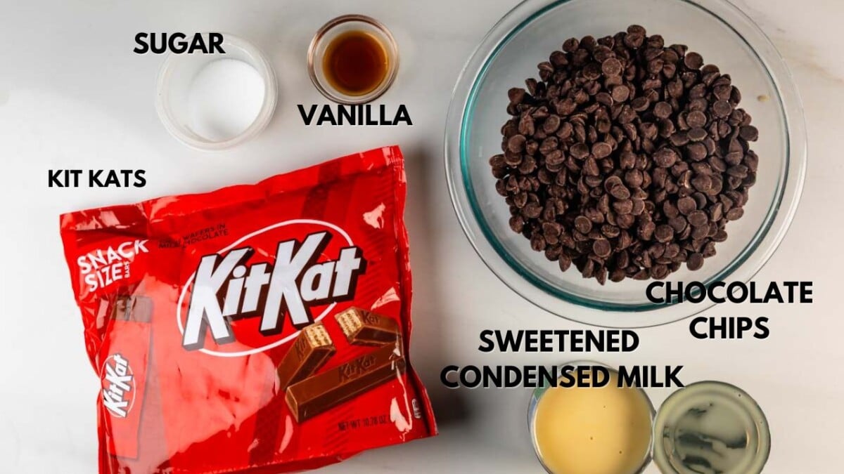 A bowl of chocolate chips, a can of sweetened condensed milk, a bowl of sugar, a small bowl of vanilla extract, and a bag of Kit Kat bars are arranged on a white surface. Each item is labeled.