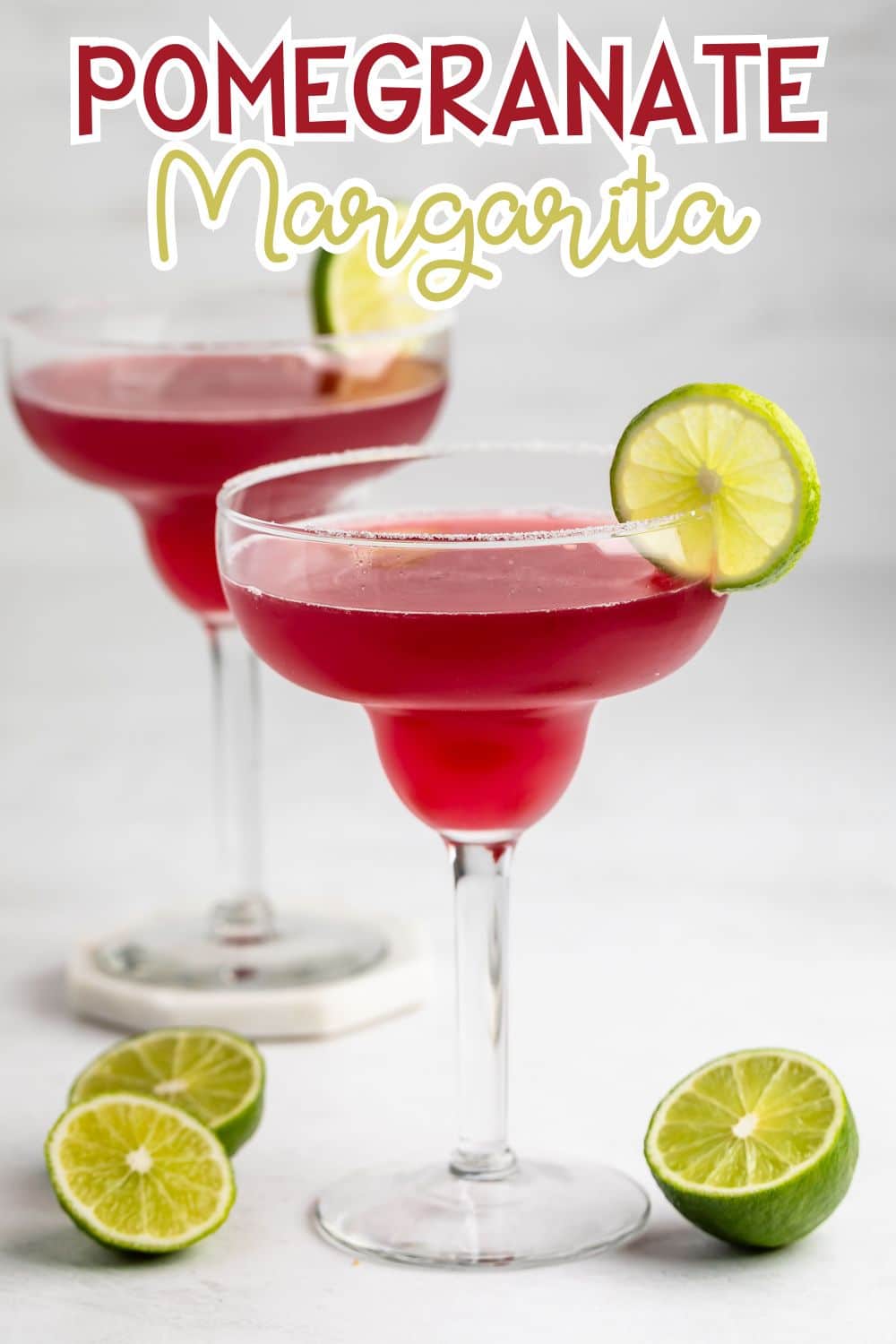 Two pomegranate margaritas in cocktail glasses with salted rims, each garnished with a lime slice. Fresh, halved limes sit on the white surface nearby. Text above reads Pomegranate Margarita.
