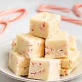 A stack of creamy white fudge squares with bits of red peppermint on a white plate. In the background, blurred candy canes are visible on a light-colored surface.
