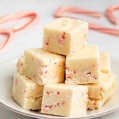 A stack of creamy white fudge squares with bits of red peppermint on a white plate. In the background, blurred candy canes are visible on a light-colored surface.