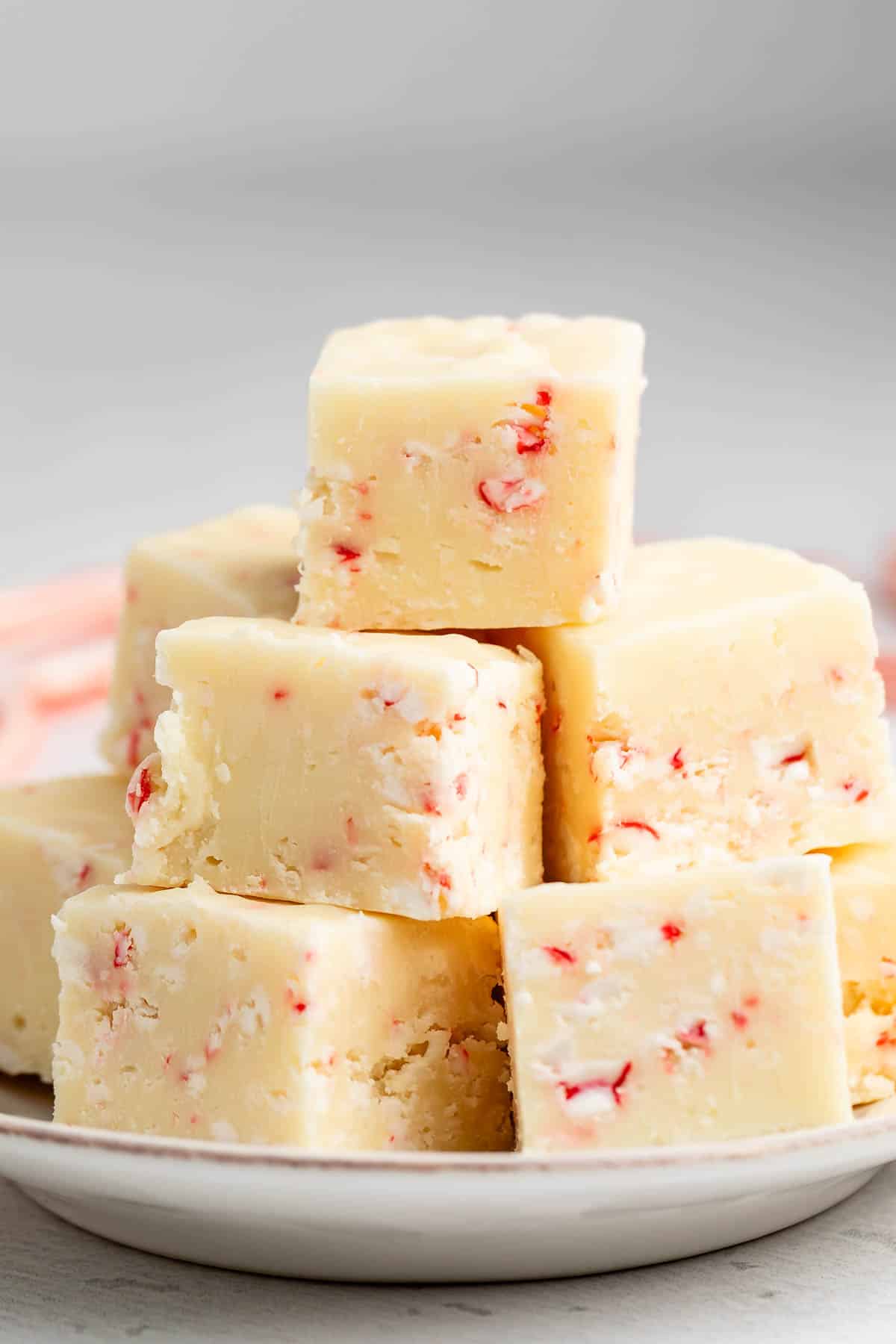 A plate stacked with creamy white fudge squares containing small red peppermint candy pieces. The fudge looks smooth and festive, with a soft texture and slightly uneven edges.