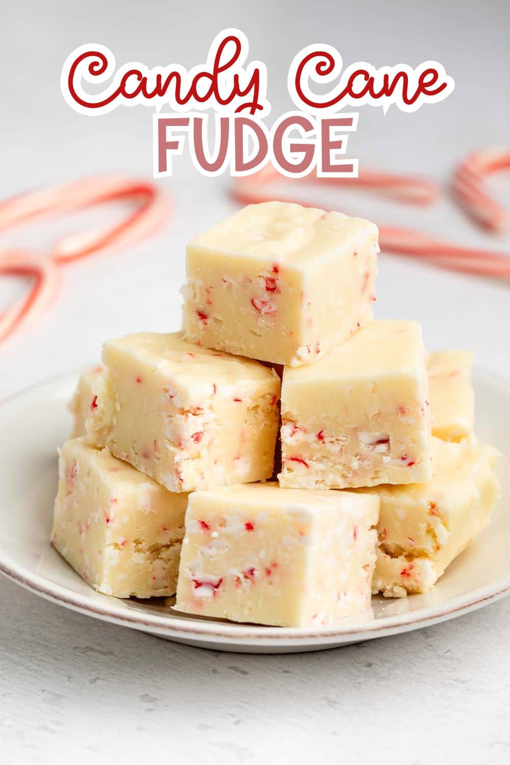 A plate stacked with creamy white fudge squares containing red candy cane pieces, with two candy canes and the text โCandy Cane Fudgeโ in bold lettering above the plate.