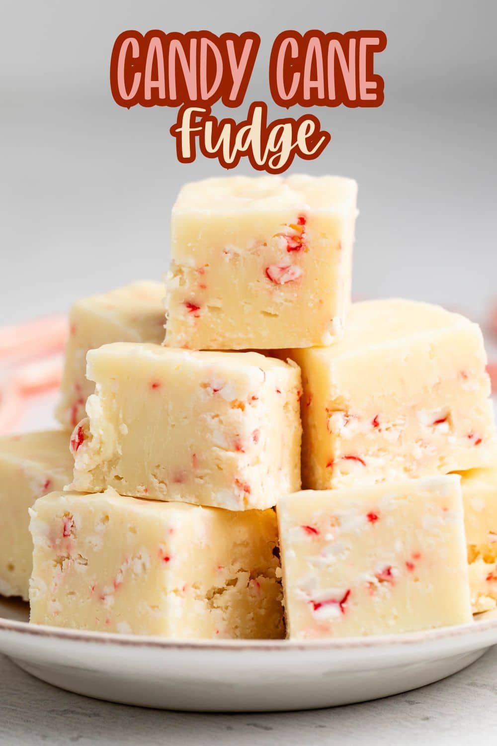 A plate stacked with creamy white fudge pieces containing red and white candy cane bits. Overhead text reads Candy Cane Fudge in bold red and brown letters.