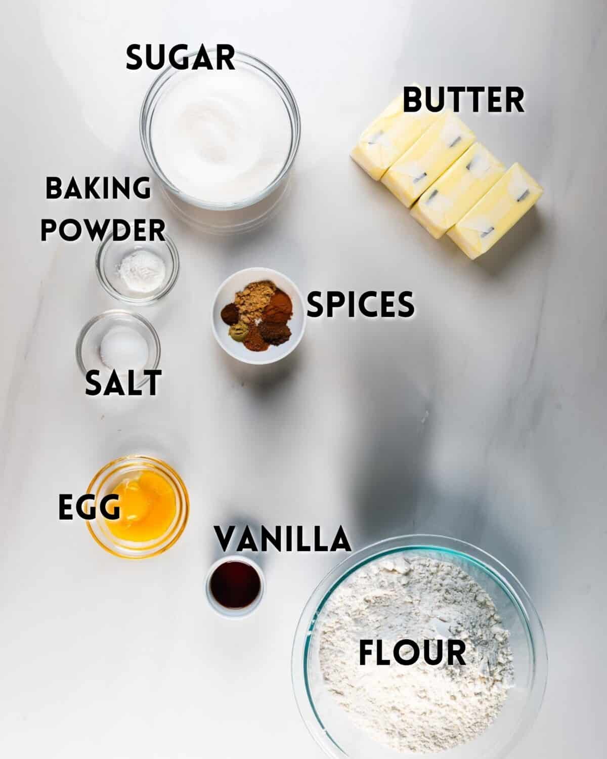 Top-down view of labeled baking ingredients on a white surface: sugar, butter sticks, flour, vanilla extract, egg, salt, baking powder, and a small bowl of assorted spices.