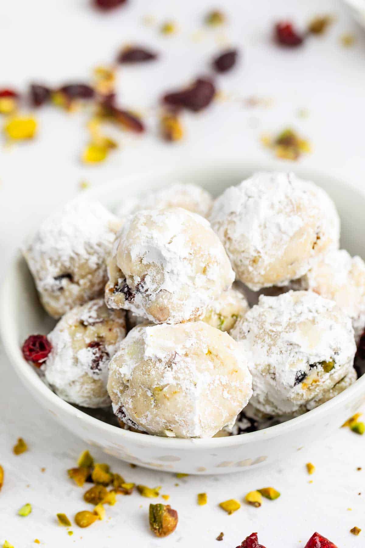 A white bowl filled with round cookies coated in powdered sugar, with pieces of pistachio and dried cranberries scattered around on a white surface.