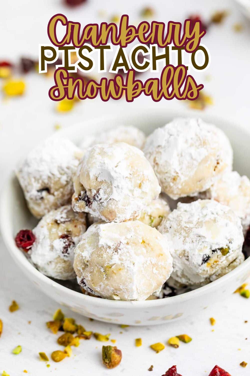 A bowl filled with round, powdered sugar-coated cranberry pistachio snowball cookies, with scattered cranberries and pistachios around, and the text Cranberry Pistachio Snowballs above.