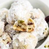 A bowl of round cookies dusted with powdered sugar, with one cookie broken in half to show a crumbly interior with pieces of pistachio and dried fruit.