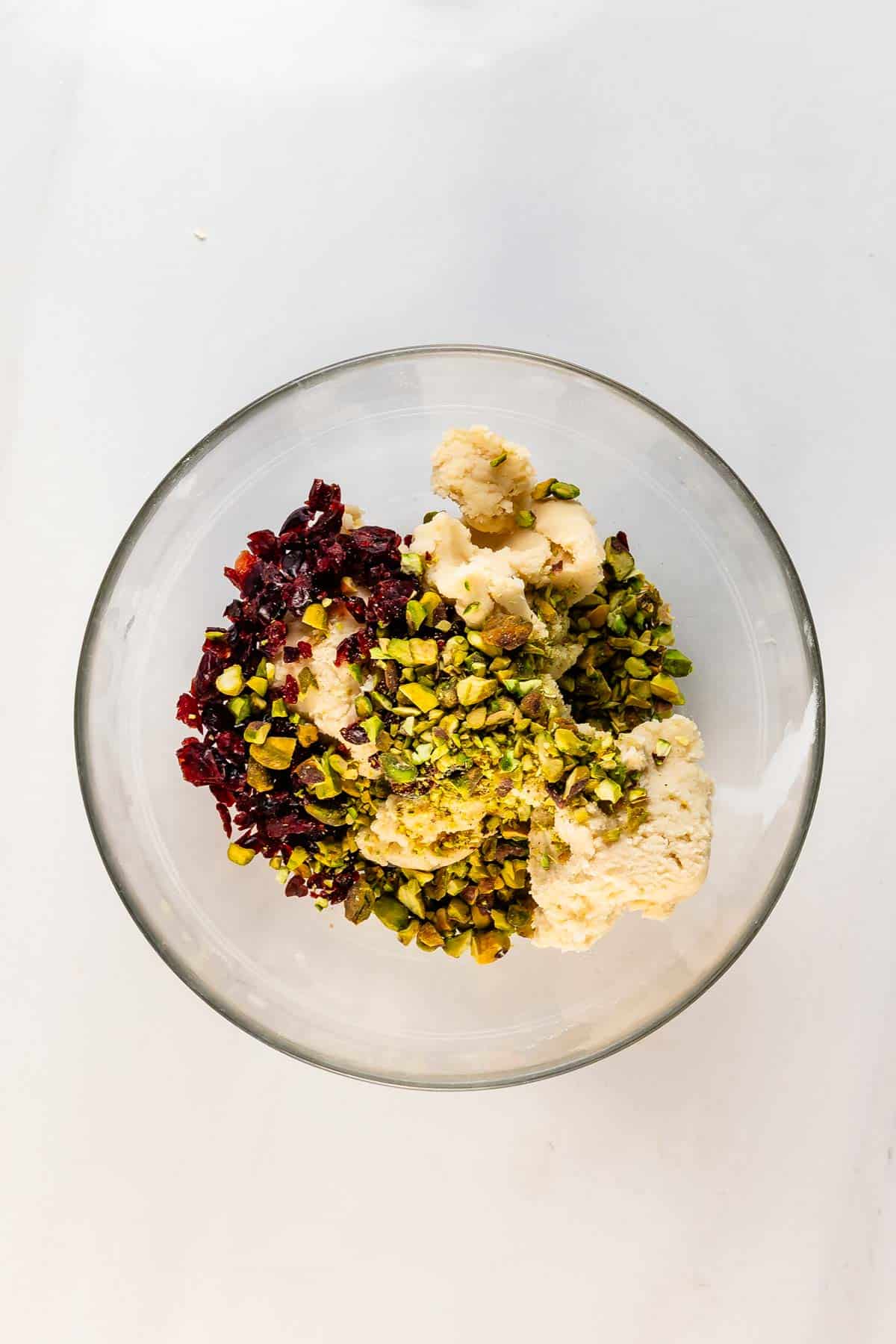 A clear glass bowl containing vanilla ice cream topped with chopped pistachios and dried cranberries, set on a white surface.