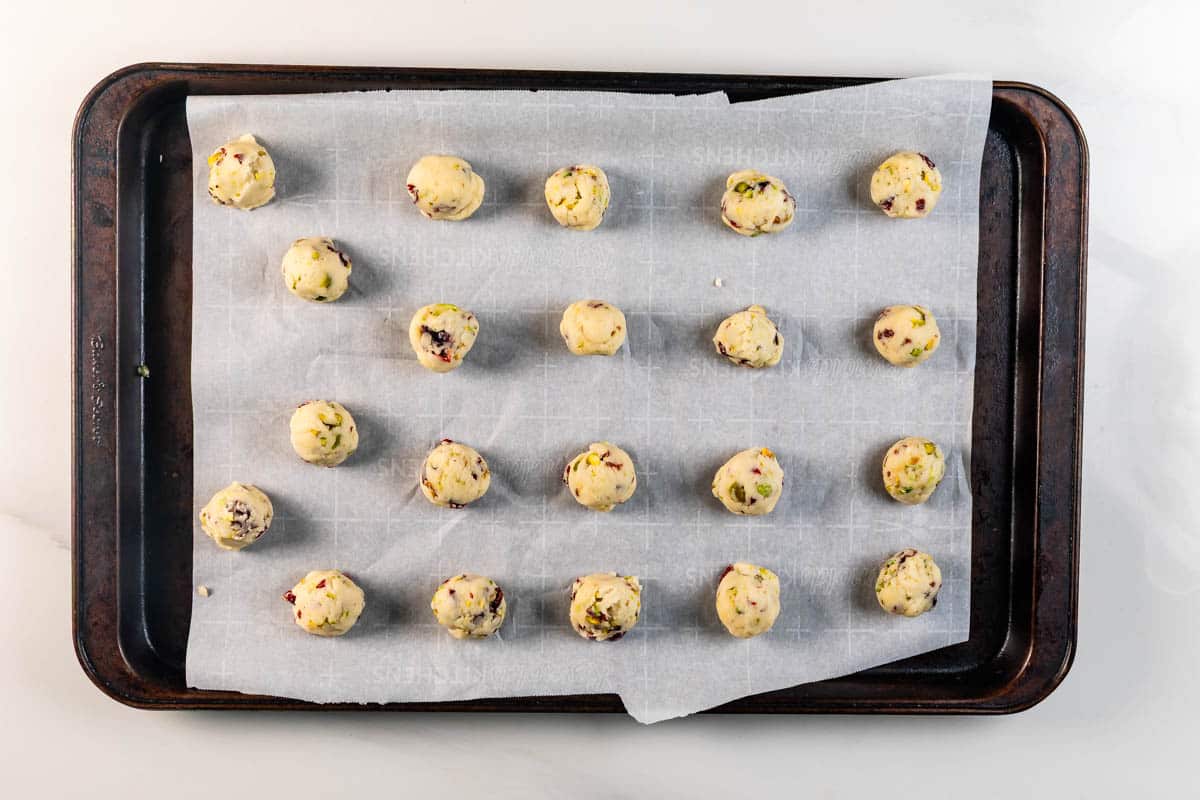 A baking tray lined with parchment paper holds 20 evenly spaced, unbaked cookie dough balls with visible chunks of nuts and fruit.