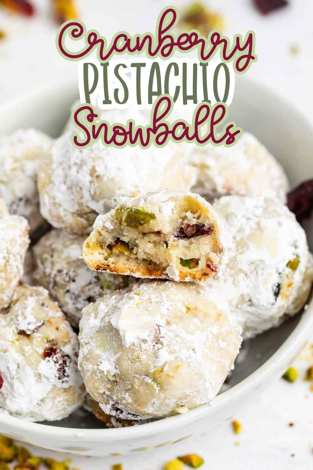 A bowl filled with cranberry pistachio snowball cookies, dusted with powdered sugar. One cookie is broken in half, showing bits of cranberry and pistachio inside. The text Cranberry Pistachio Snowballs is overlaid.