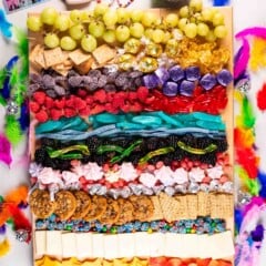 A colorful snack board with grapes, candies, cookies, crackers, cheese, and gummies, decorated with rainbow feathers, beaded bracelets, a small disco ball, and Taylor Swift concert-themed items.