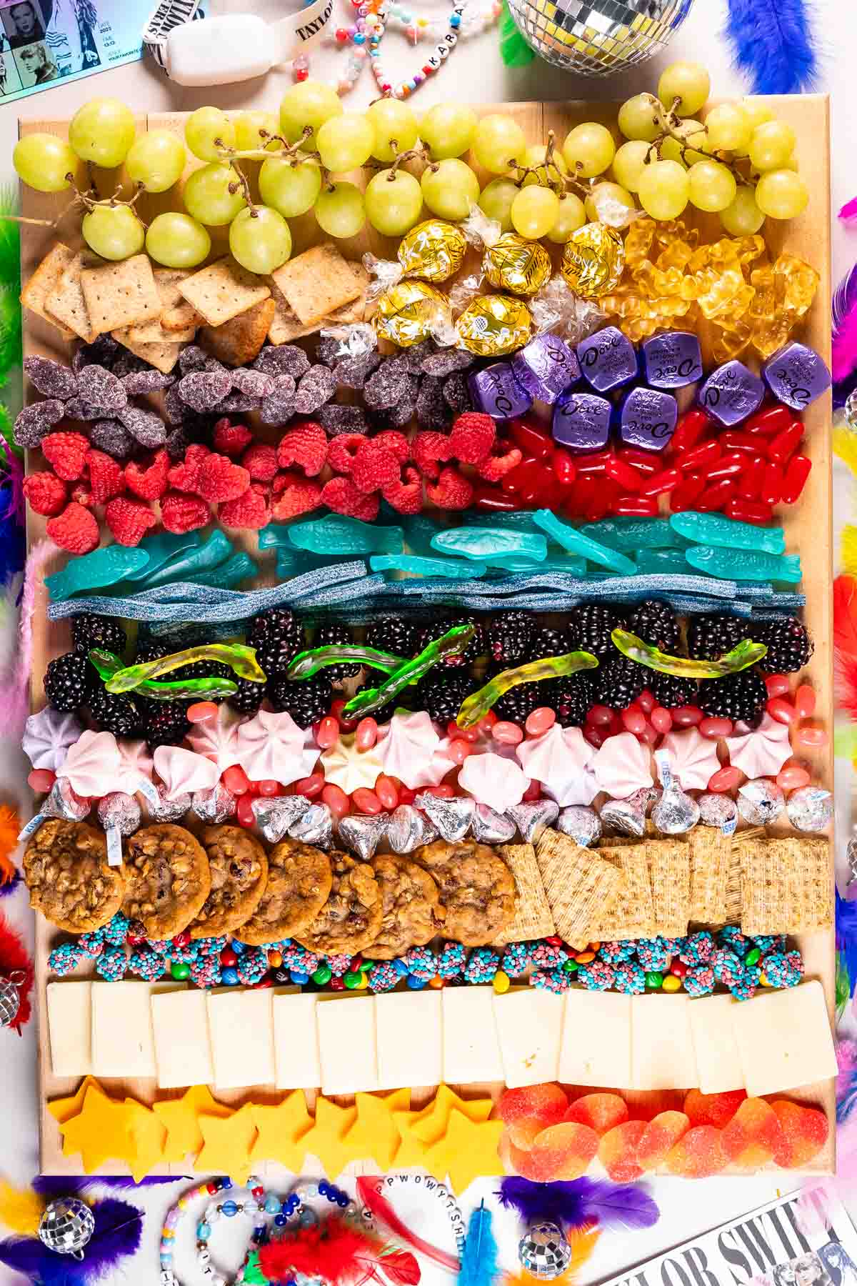 A colorful tray filled with grapes, crackers, cookies, candies, licorice, chocolates, and cheese, arranged in neat horizontal rows. The tray is surrounded by feathers, beads, and party decorations.