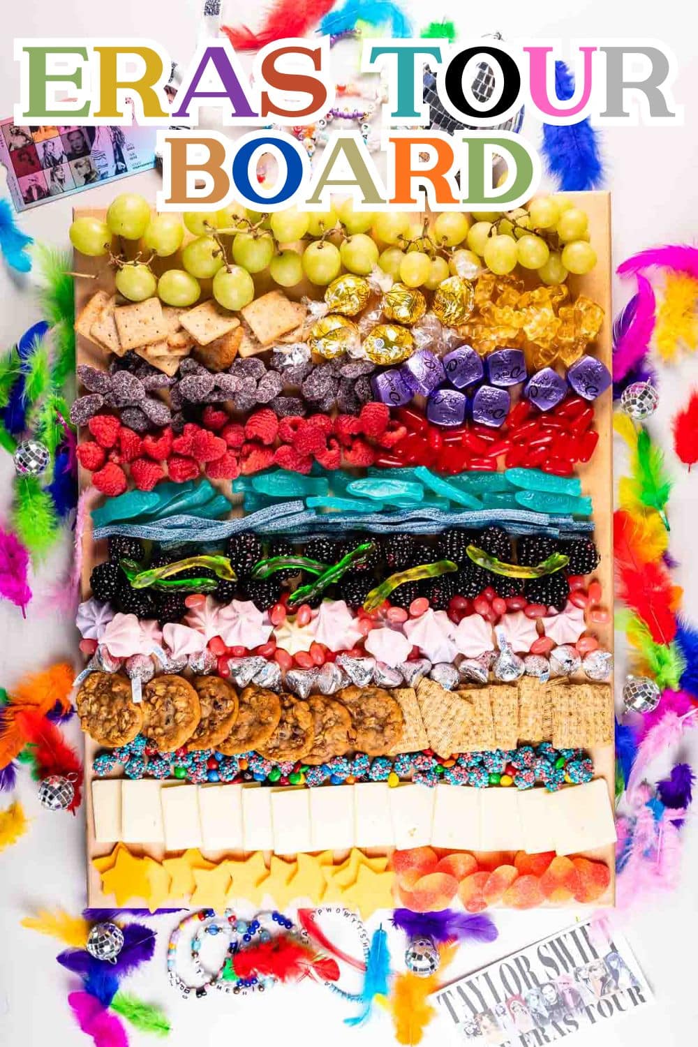 A colorful snack board inspired by Taylor Swifts Eras Tour, featuring rows of different candies, cookies, cheeses, grapes, and crackers, surrounded by bright feathers and tour-themed decorations. Text reads Eras Tour Board.