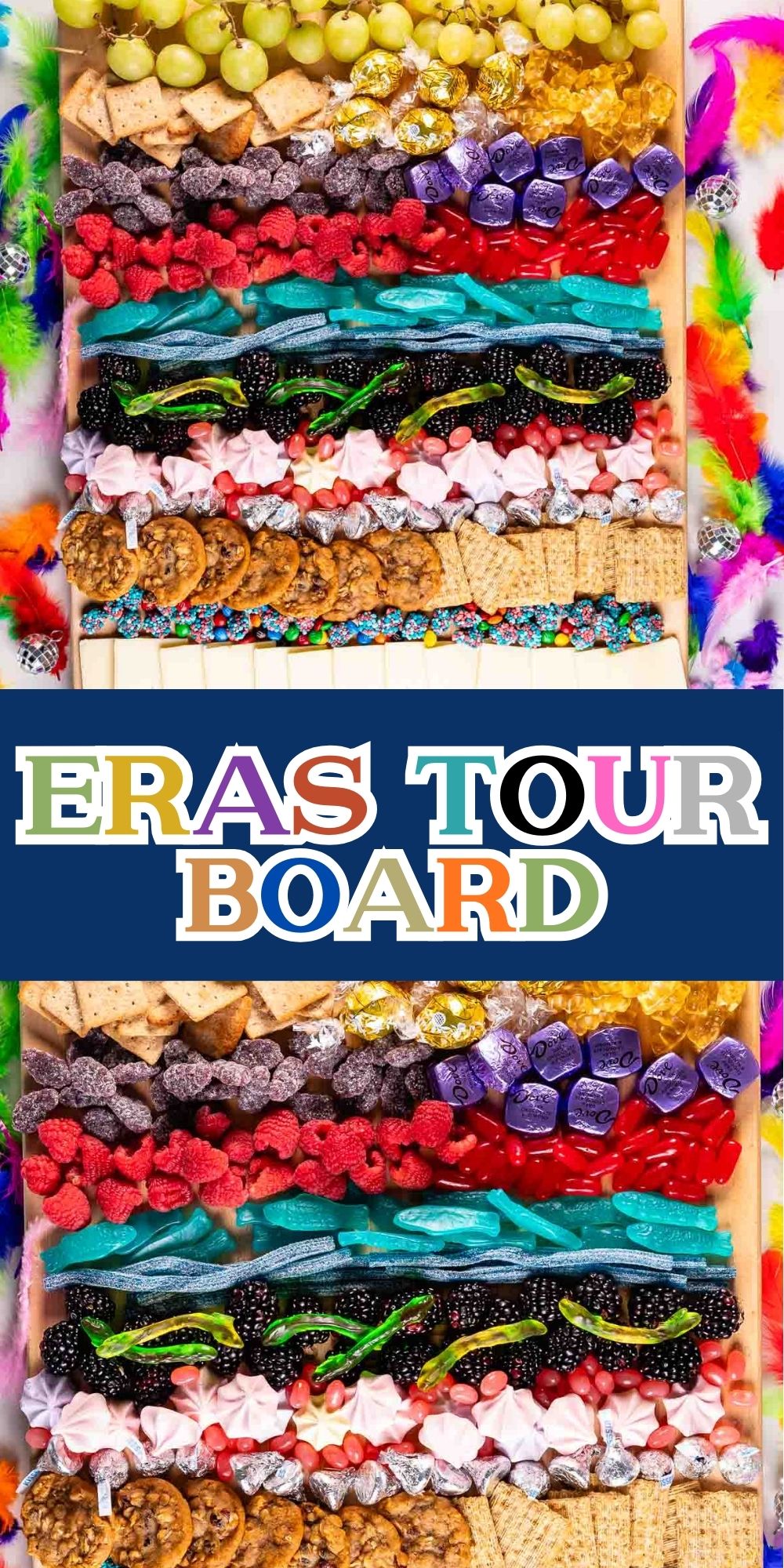 A colorful snack board filled with grapes, cheese, candies, pretzels, and crackers, decorated with vibrant feathers. In the center, text reads ERAS TOUR BOARD in multicolored letters on a navy background.