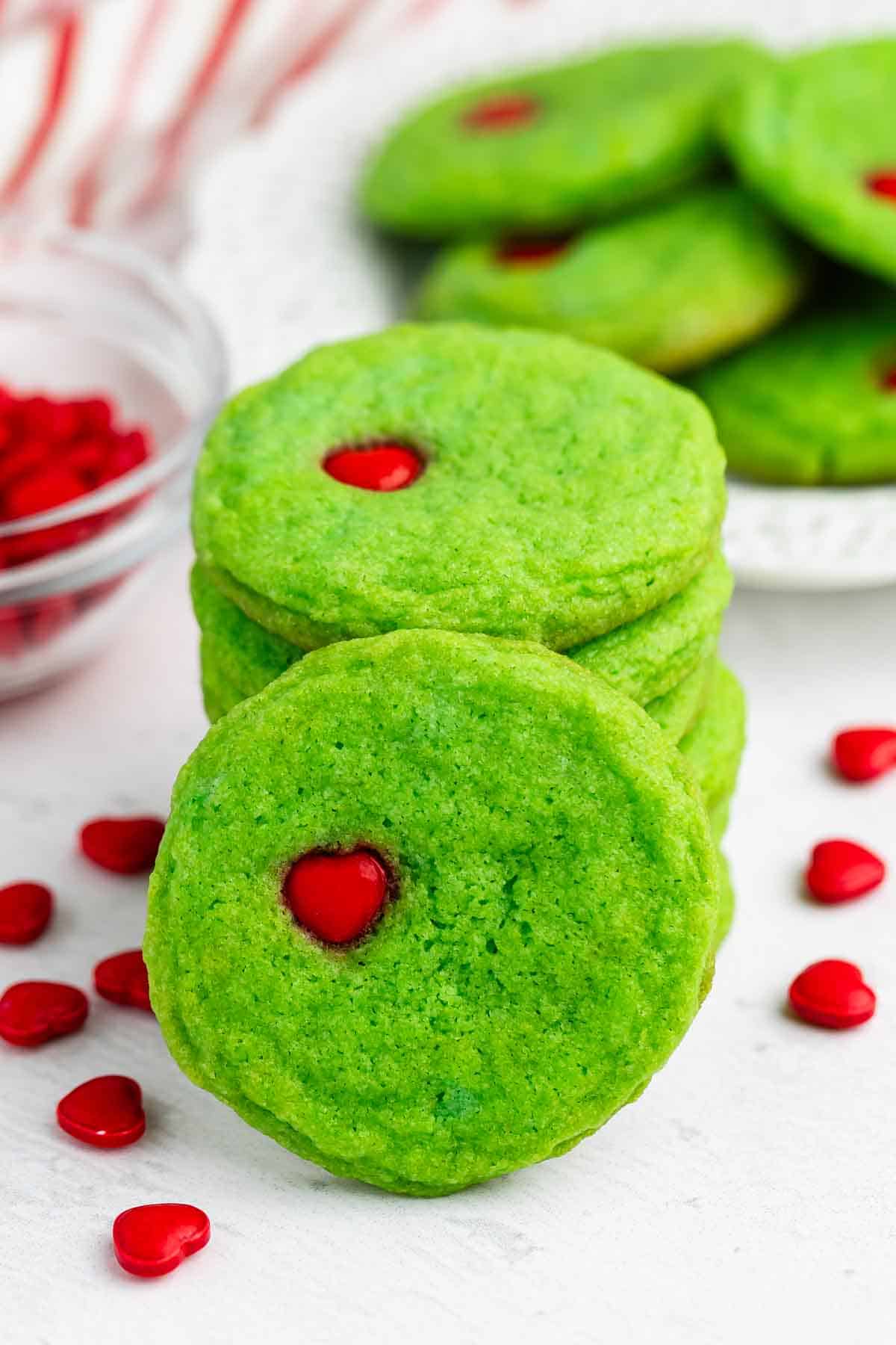 Three bright green cookies with red heart-shaped candies in the center are stacked in front of a plate with more cookies and red heart-shaped candies scattered around on a white surface.