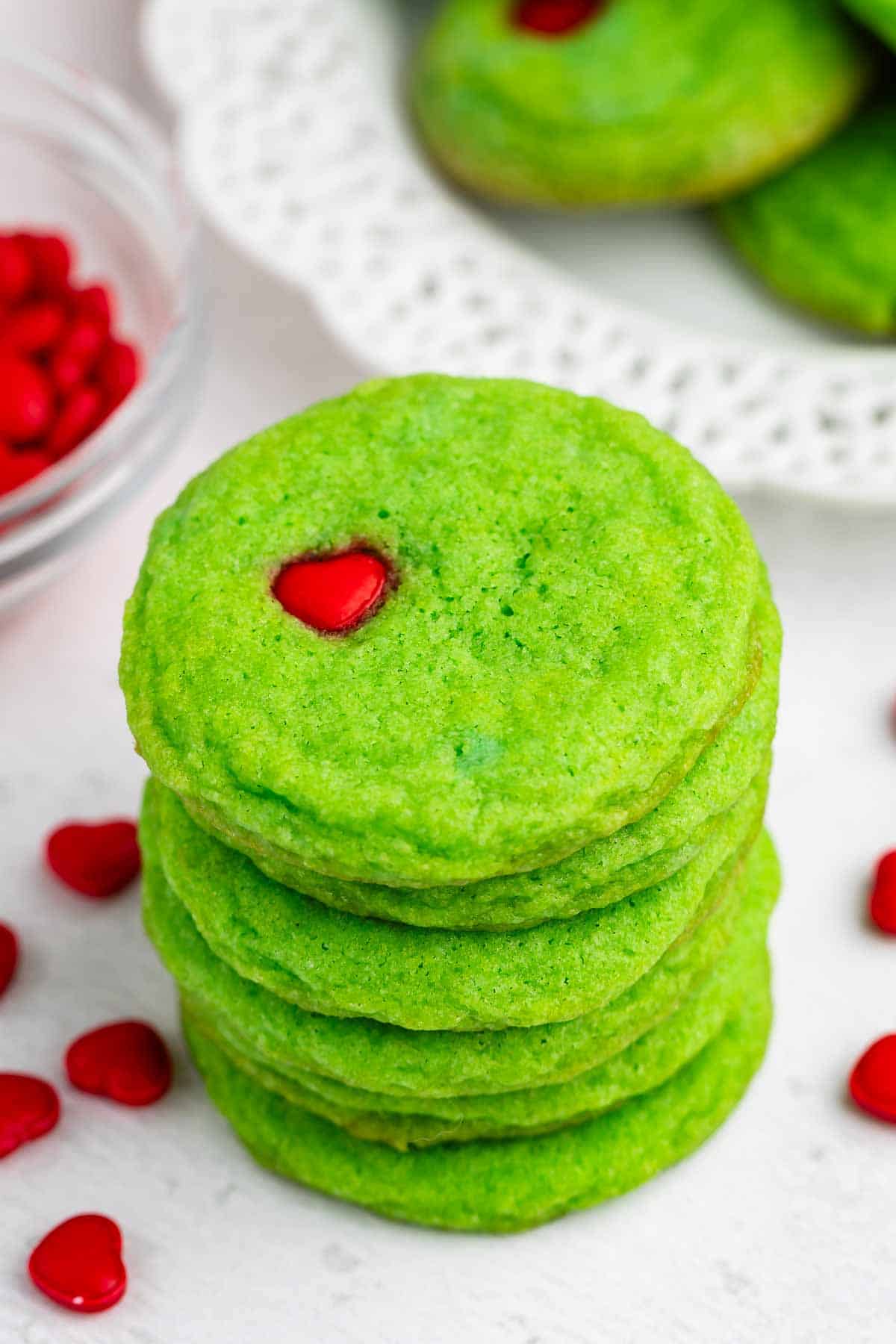 A stack of green cookies with a small red heart candy on top of the uppermost cookie. Red heart candies are scattered around, and more cookies are visible on a white plate in the background.