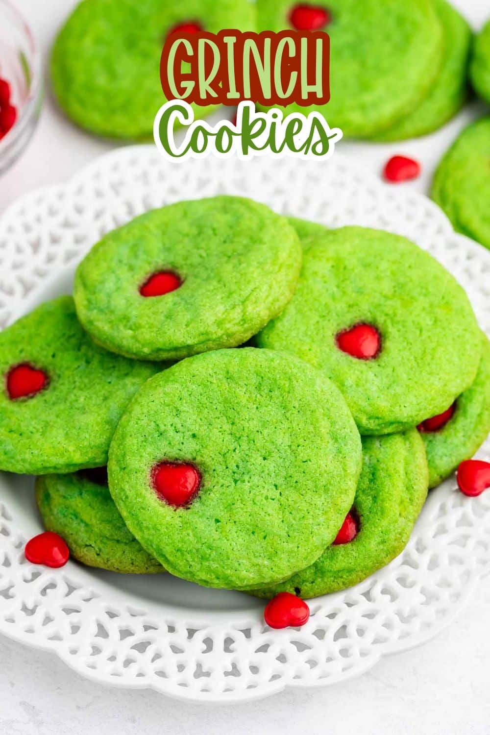 A plate of bright green cookies with red candy hearts in the center, arranged on a white decorative plate. The words Grinch Cookies appear at the top of the image.
