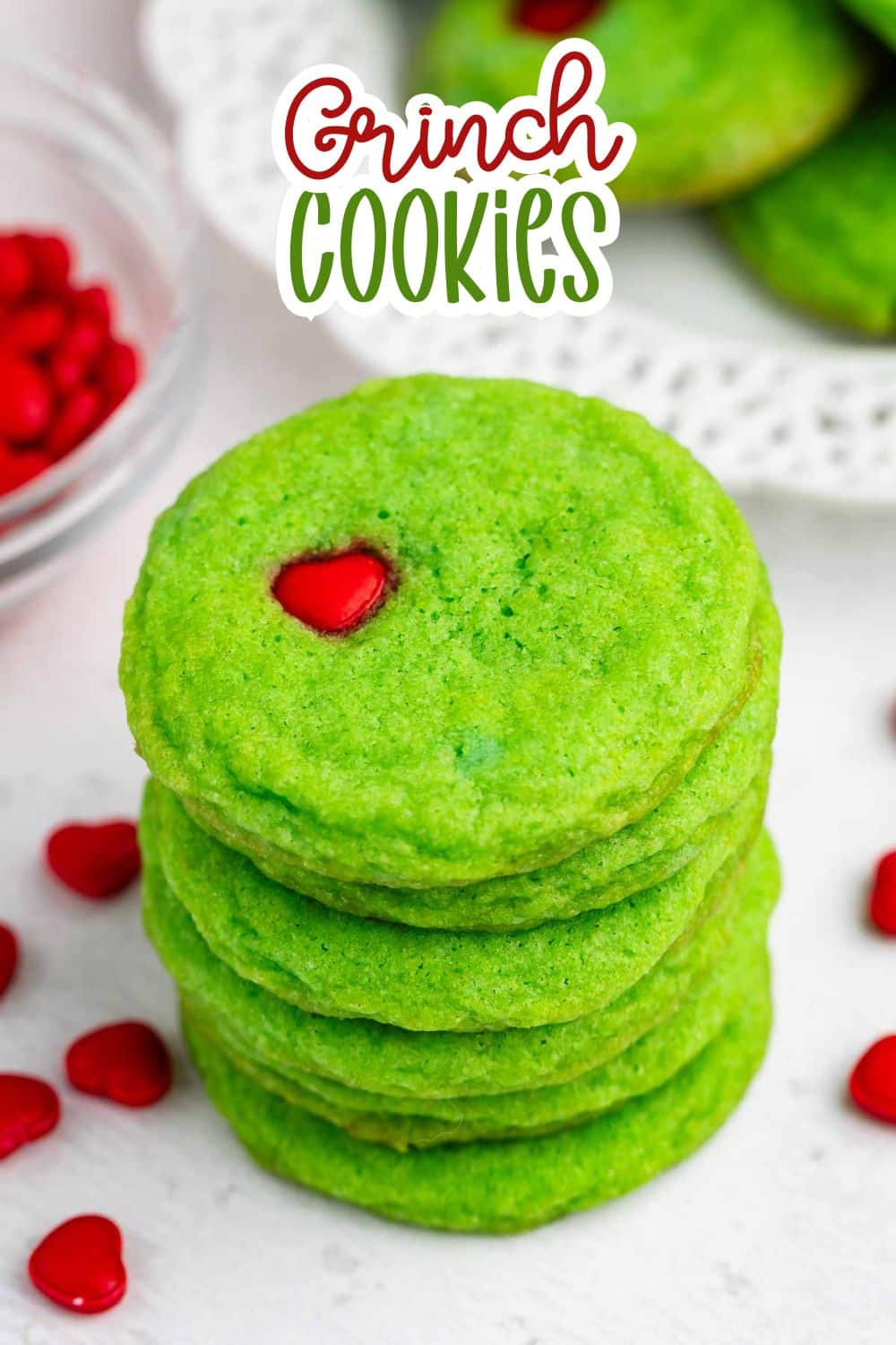 A stack of bright green cookies, each topped with a single red heart-shaped candy, sits on a white surface. The text “Grinch Cookies” appears above the cookies. Red candies are scattered nearby.