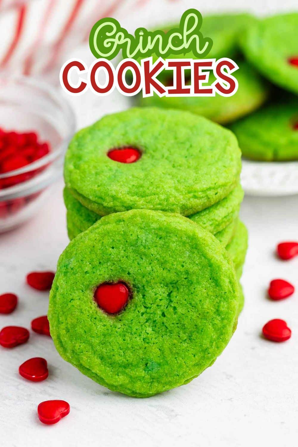 A stack of bright green cookies with red heart-shaped candies in the center. More cookies and red heart candies are scattered nearby. The text Grinch Cookies is at the top of the image.