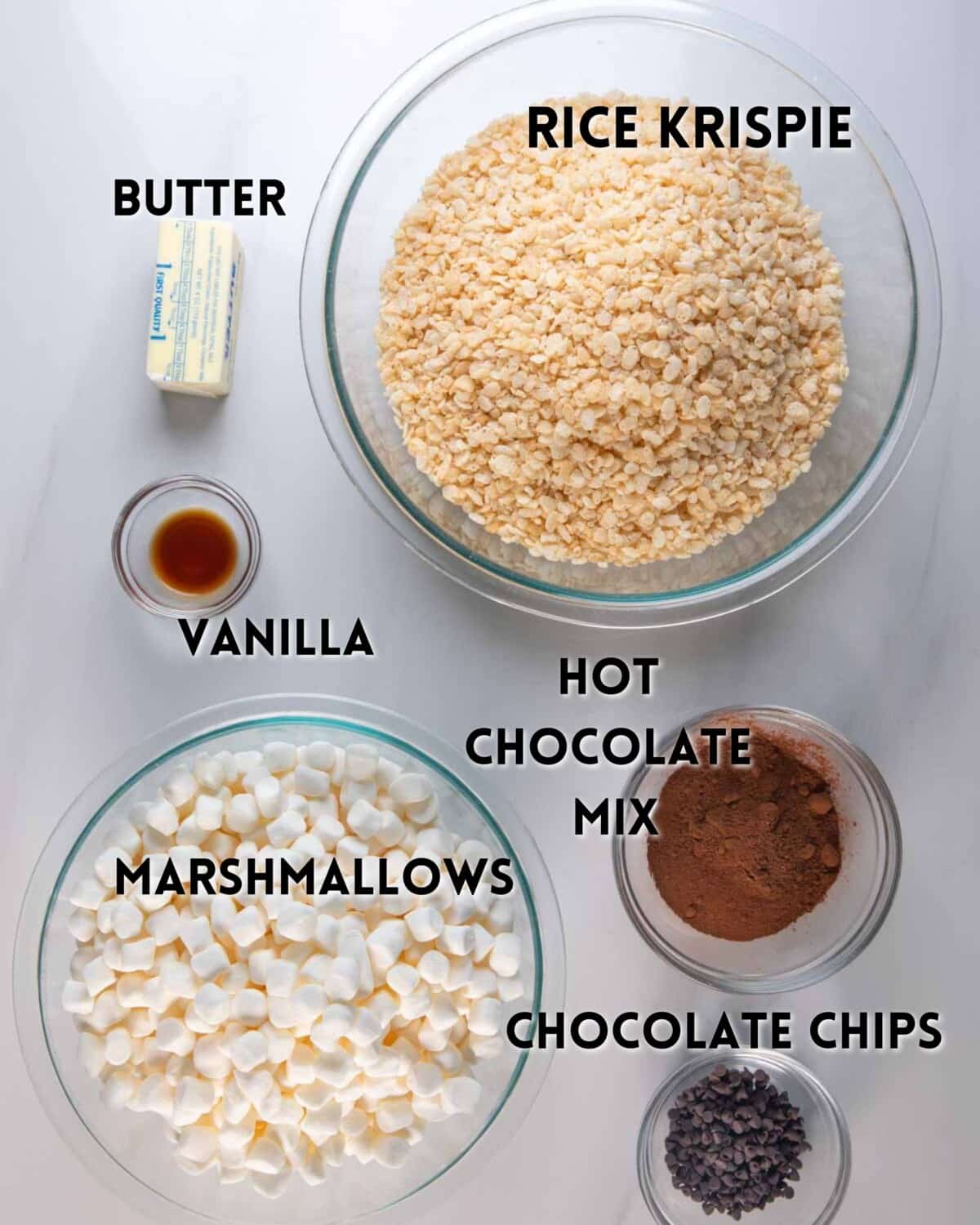 A top-down view of labeled baking ingredients in bowls: rice krispies, mini marshmallows, chocolate chips, hot chocolate mix, a stick of butter, and vanilla extract on a light surface.
