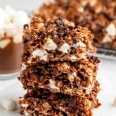 A stack of three chocolate crispy rice treats with mini marshmallows and chocolate chips, with more treats and a mug of hot chocolate with marshmallows in the background.