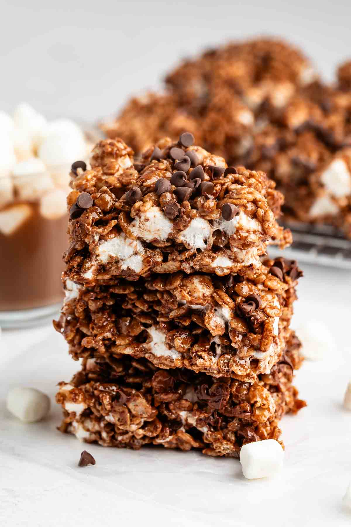 A stack of three chocolate crispy rice treats with mini marshmallows and chocolate chips, with more treats and a mug of hot chocolate with marshmallows in the background.