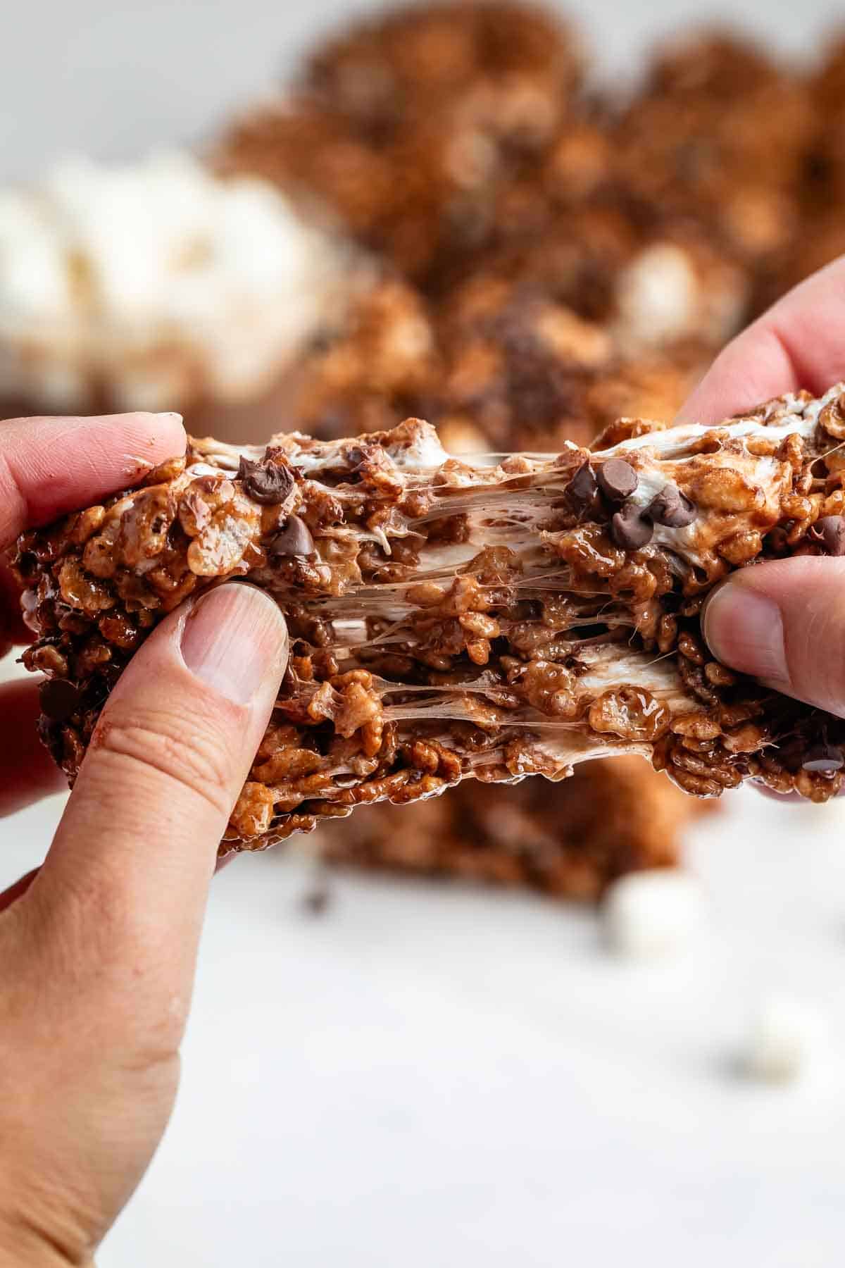 Close-up of hands pulling apart a gooey chocolate rice crispy treat, showing melted marshmallow stretching between the two pieces. The background is blurred with more treats and marshmallows visible.