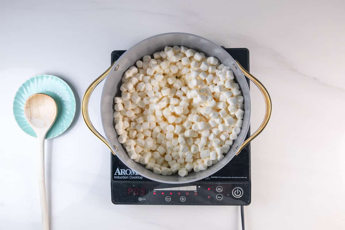 A pot filled with mini marshmallows sits on an induction cooktop. Next to it is a turquoise plate holding a wooden spoon, all on a white countertop.