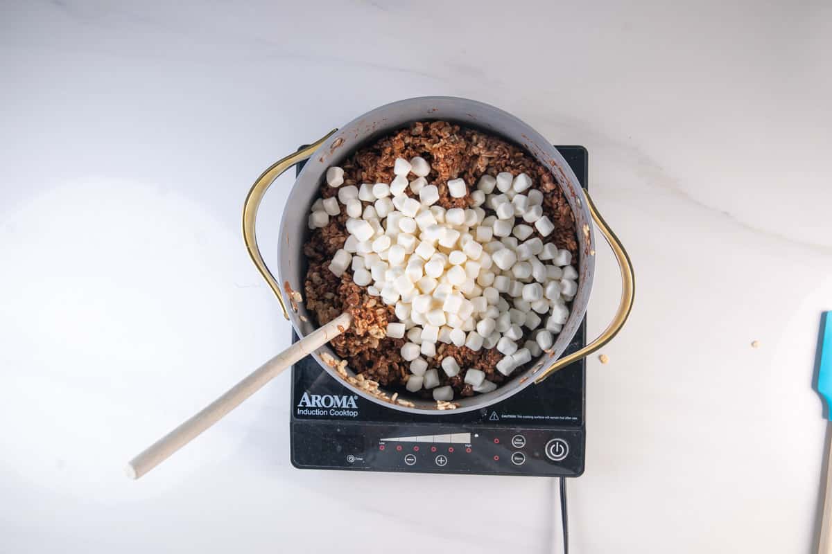 A pot on an electric cooktop contains chocolatey, crispy cereal mixed with mini marshmallows, stirred with a wooden spoon. The scene is set on a white countertop.
