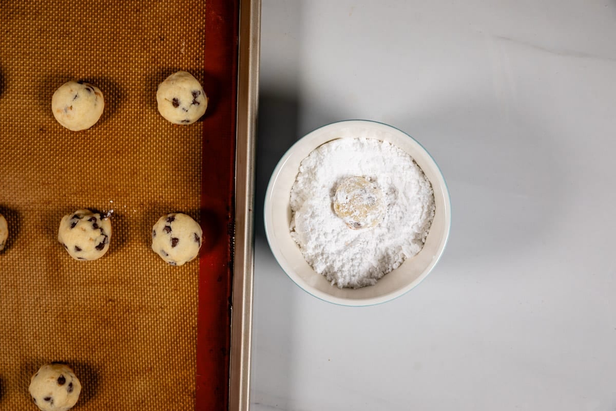 A bowl of powdered sugar with a cookie dough ball inside sits next to a baking sheet lined with silicone, holding several chocolate chip cookie dough balls.