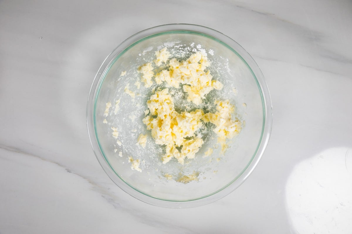 A glass mixing bowl containing partially creamed butter and sugar sits on a white marble surface.