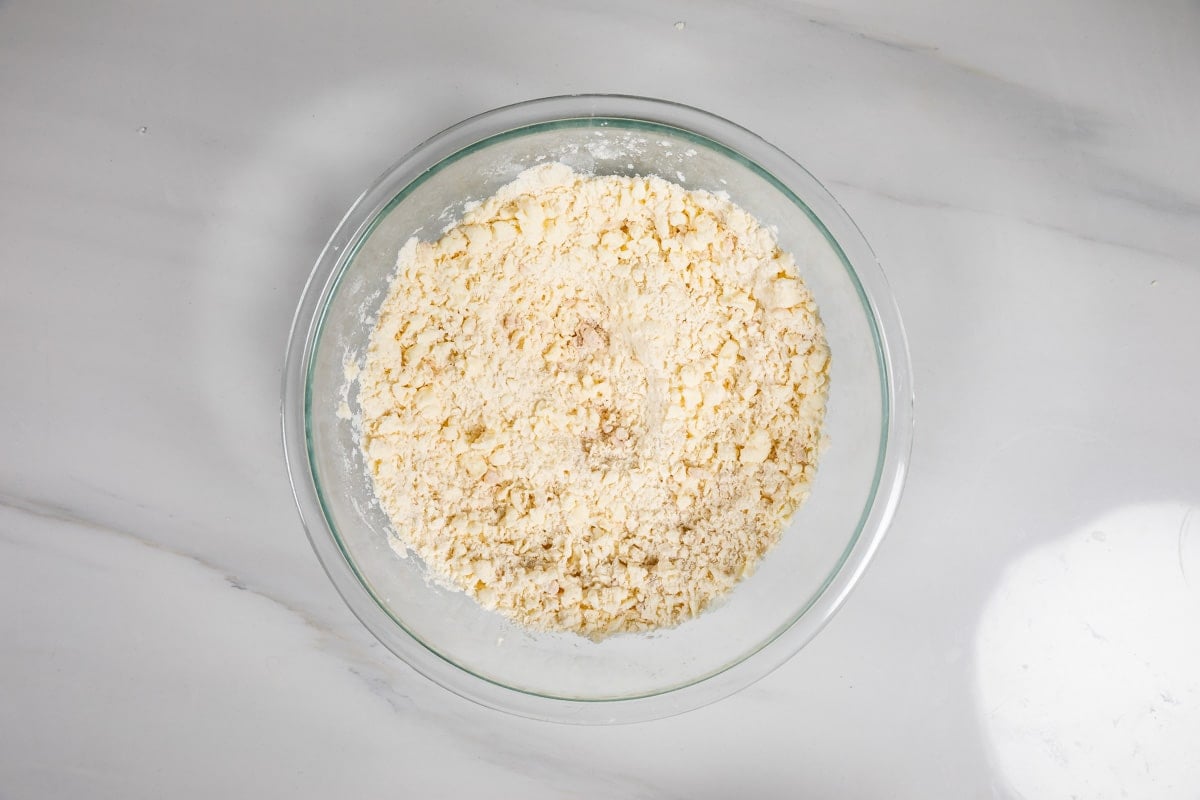 A glass bowl filled with crumbly, pale yellow dough mixture sits on a white marble surface.