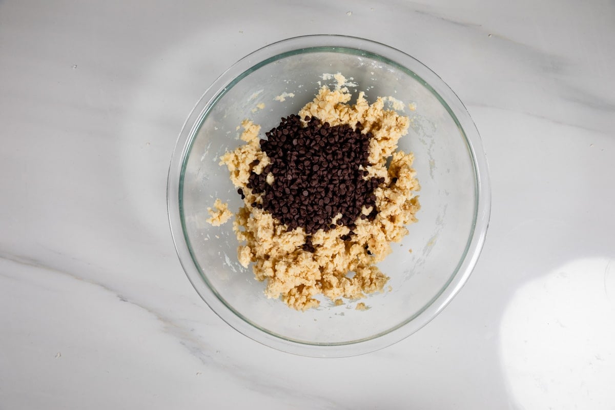 A glass bowl on a white countertop contains cookie dough with a pile of chocolate chips in the center, ready to be mixed.