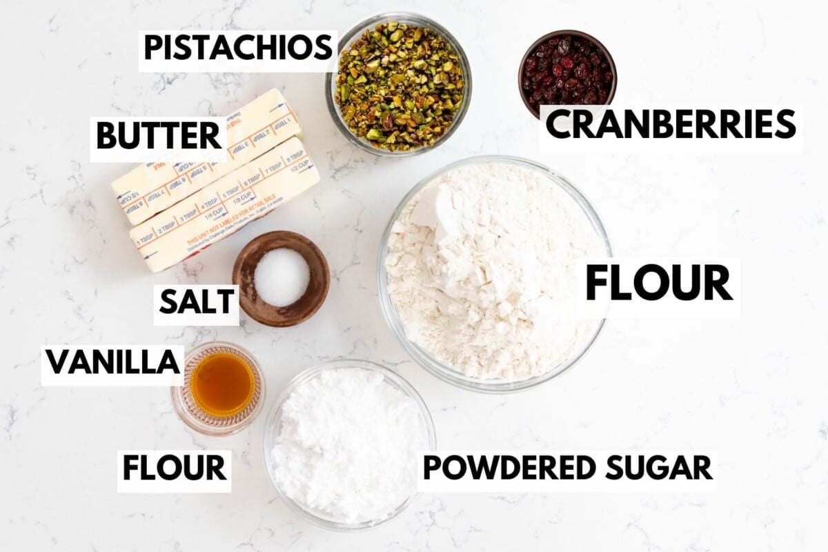 Baking ingredients on a white surface: butter sticks, chopped pistachios, dried cranberries, flour, salt in a small bowl, vanilla extract, and powdered sugar, each labeled with bold text.