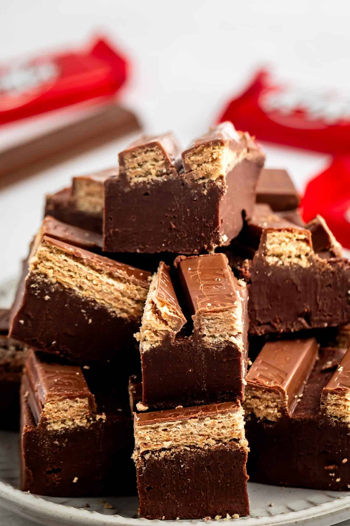 A stack of chocolate-covered wafer bars, some broken to show crispy wafer layers inside, sits on a white plate with red-wrapped bars blurred in the background.