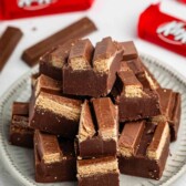 A plate stacked with cut pieces of chocolate wafer bars, revealing layers of chocolate and wafer inside. Red wrappers with KitKat branding are visible in the background.