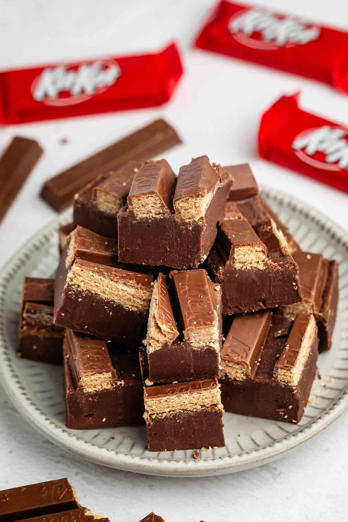 A plate stacked with cut pieces of chocolate wafer bars, revealing layers of chocolate and wafer inside. Red wrappers with KitKat branding are visible in the background.