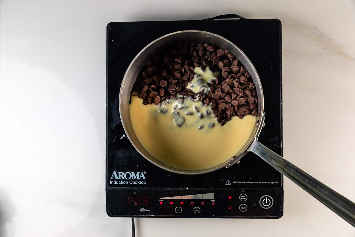 A saucepan on an Aroma induction cooktop contains chocolate chips and sweetened condensed milk, starting to melt together.