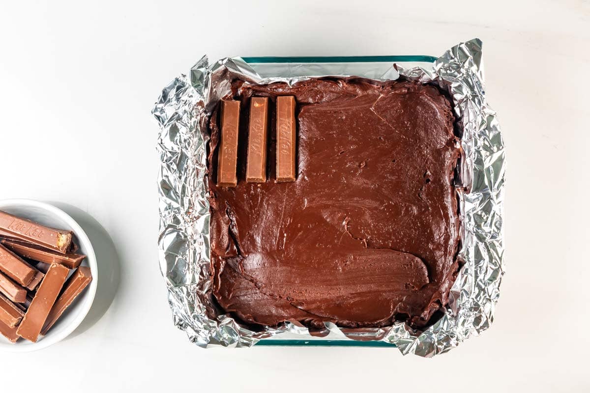 A square pan lined with foil holds chocolate fudge, topped with three Kit Kat bars in one corner. Next to it is a small bowl filled with additional Kit Kat bars.