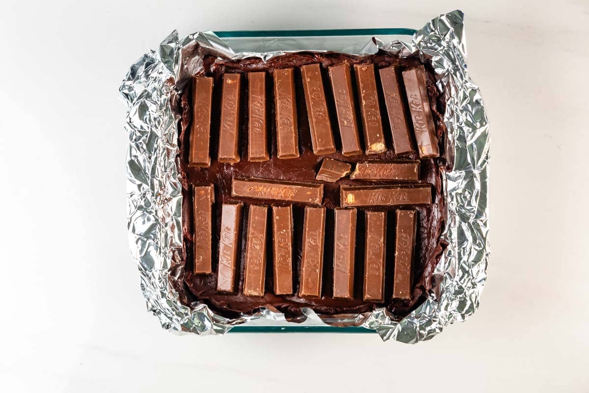 A baking dish lined with foil holds brownie batter topped with rows of Kit Kat chocolate bars, ready to be baked.