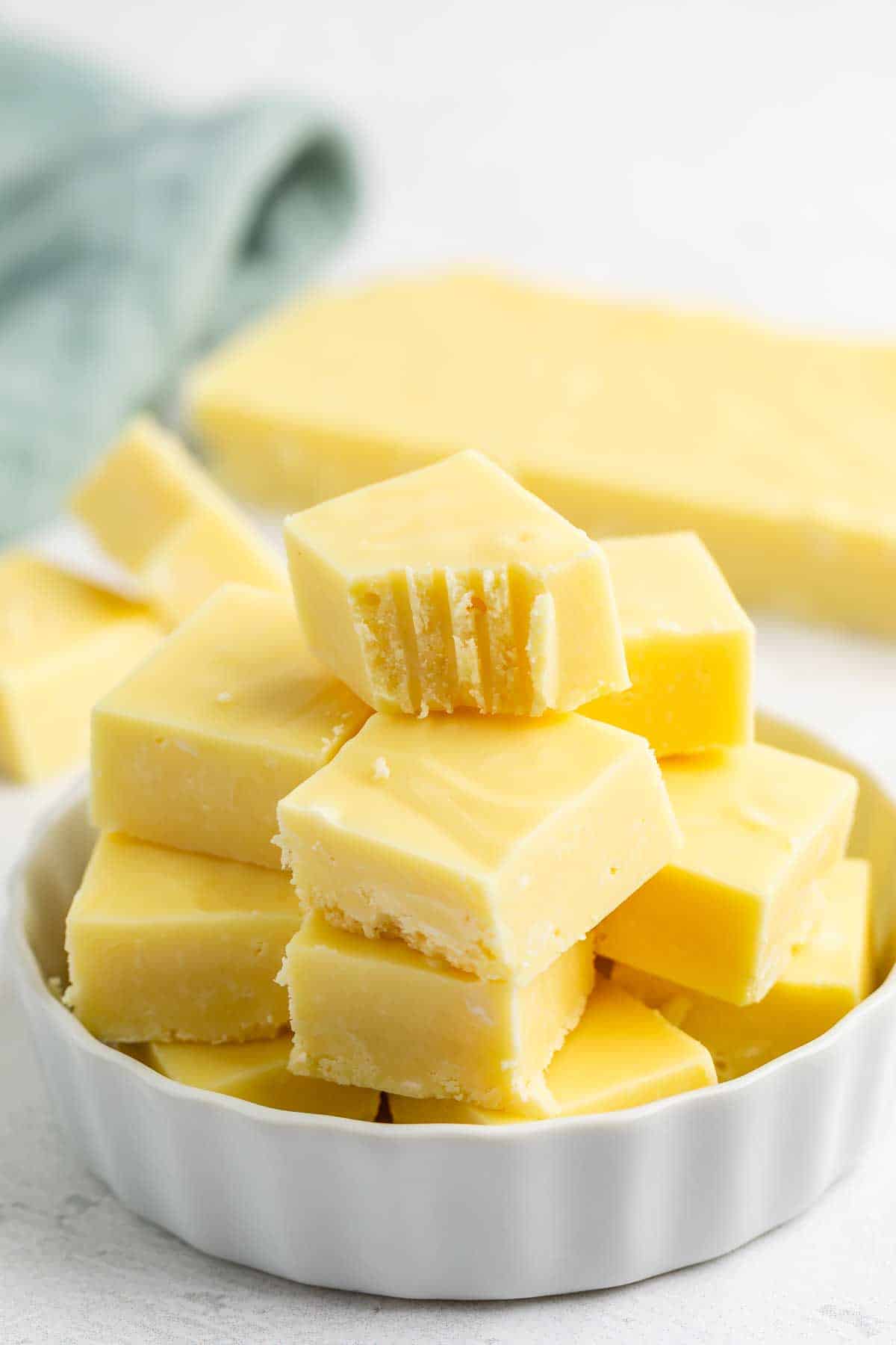 A stack of creamy yellow fudge squares is arranged on a white dish. Some pieces have bite marks. A block of fudge and a blue cloth are blurred in the background.