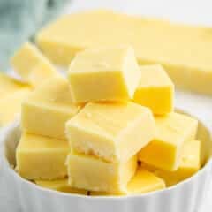 A white dish filled with neatly stacked pieces of creamy yellow fudge, with more fudge and a green cloth blurred in the background.