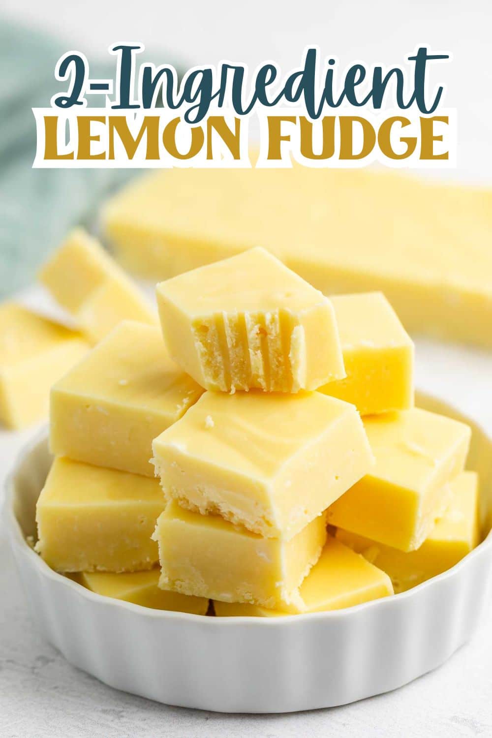 A white dish filled with yellow lemon fudge squares, some with bite marks. Text above reads “2-Ingredient Lemon Fudge.” More fudge is blurred in the background.