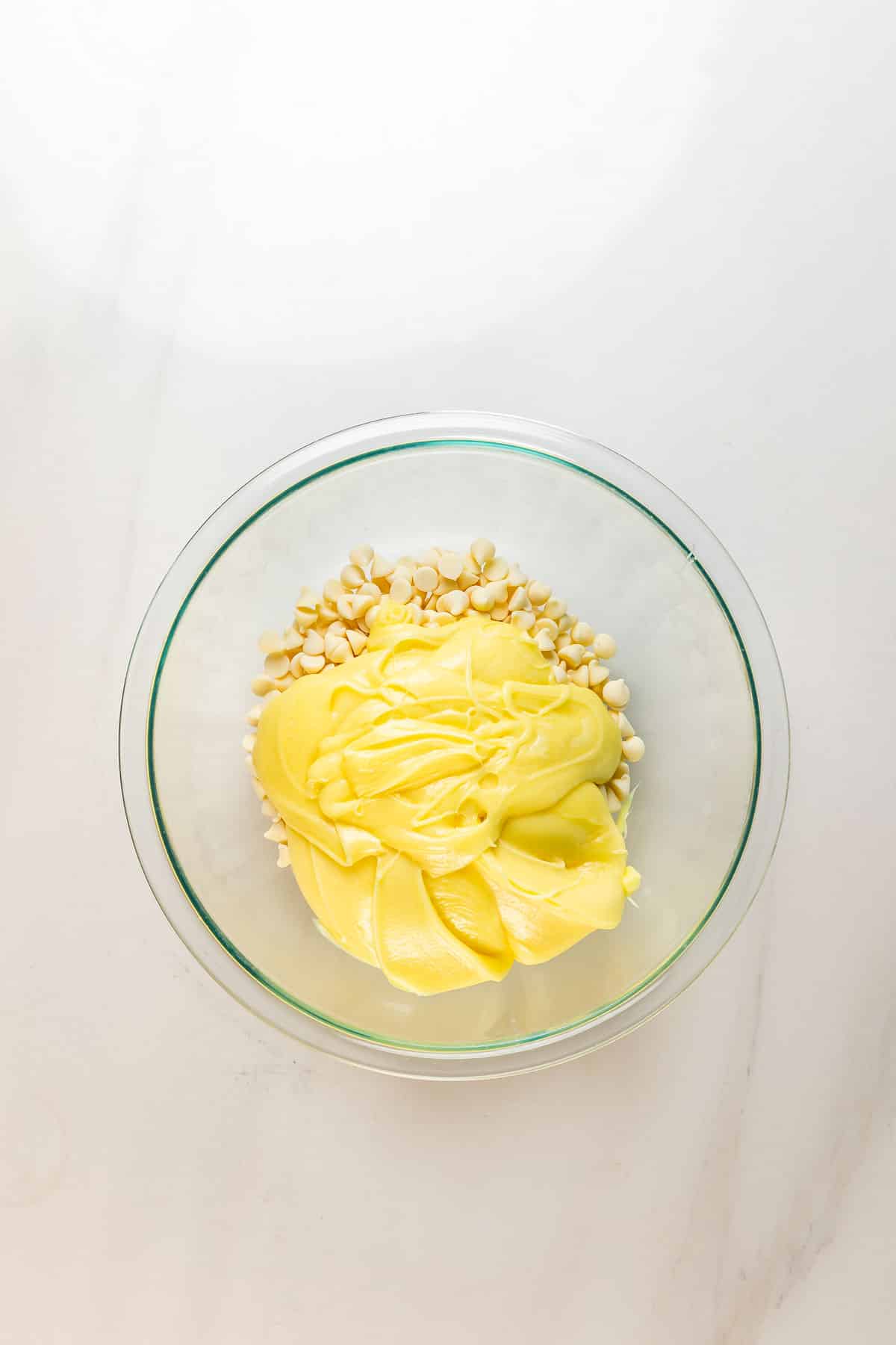 A glass bowl on a white surface containing a pile of white chocolate chips topped with a thick layer of yellow melted butter or candy coating.