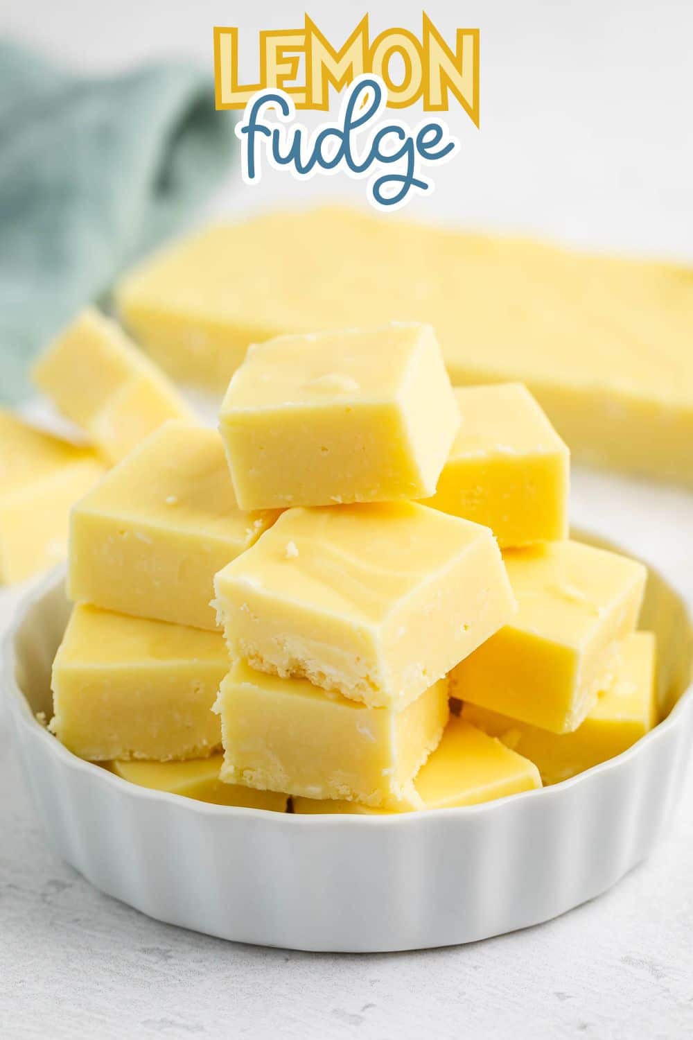 A white dish filled with yellow lemon fudge squares stacked in a pyramid shape, with more fudge and a green cloth in the blurred background. The words Lemon fudge appear at the top of the image.