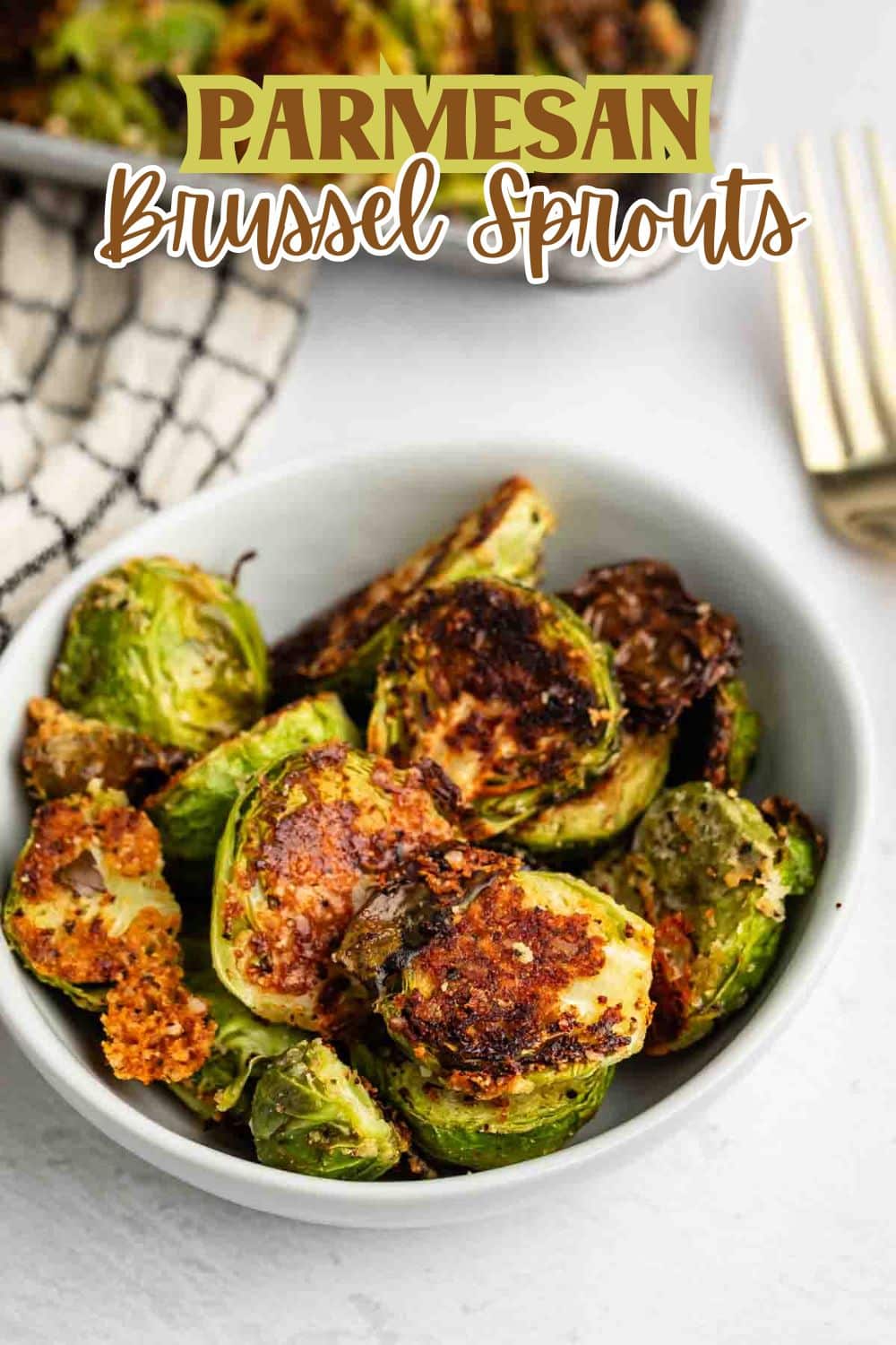 A white bowl filled with crispy roasted Brussels sprouts topped with golden, melted Parmesan cheese. A fork and a checkered towel are nearby. Text above reads Parmesan Brussel Sprouts.