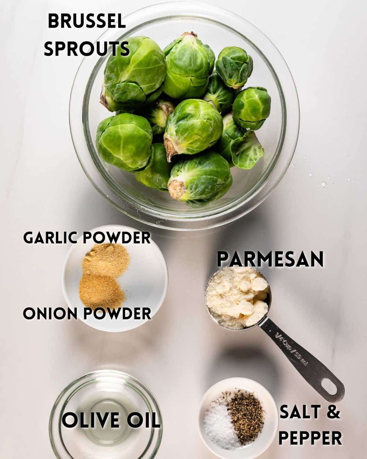A flat lay of ingredients for a Brussels sprouts dish: a bowl of Brussels sprouts, a small bowl of garlic powder, a small bowl of onion powder, a cup of grated parmesan, a bowl of olive oil, and a bowl of salt and pepper.