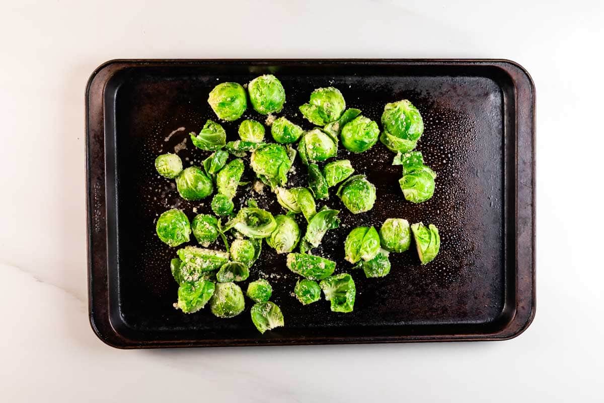 A baking tray with halved Brussels sprouts sprinkled with grated cheese and seasoning, ready to be roasted. The tray sits on a white countertop.